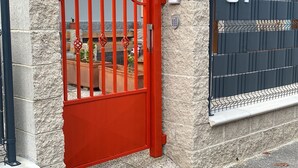 Un ingresso recintato con cancello rosso, un muro in pietra e un balcone con ringhiera metallica.