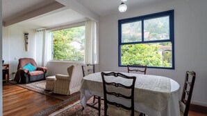 A dining area with a table set for four, surrounded by chairs, a sofa, and a window with a view of greenery. - Piscina Lareira e Natureza Incr vel - Petr Polis (Petrópolis)