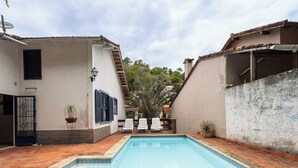 A swimming pool with a tiled deck, a white building with blue shutters, and a door with a metal gate. - Piscina Lareira e Natureza Incr vel - Petr Polis (Petrópolis)