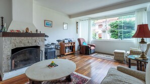 A living room with a stone fireplace, a drum set, a bookshelf, a comfortable armchair, a round coffee table, and a view of a building through a large window. - Piscina Lareira e Natureza Incr vel - Petr Polis (Petrópolis)