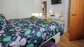 A bedroom with a bed, a bedside table with a lamp, a mirror, and a TV on the wall. - Drapers Cottage (Grange-over-Sands)