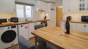 A compact kitchen with white cabinets, a wooden countertop, a built-in washing machine, and a dining area with a table and chairs. - Drapers Cottage (Grange-over-Sands)