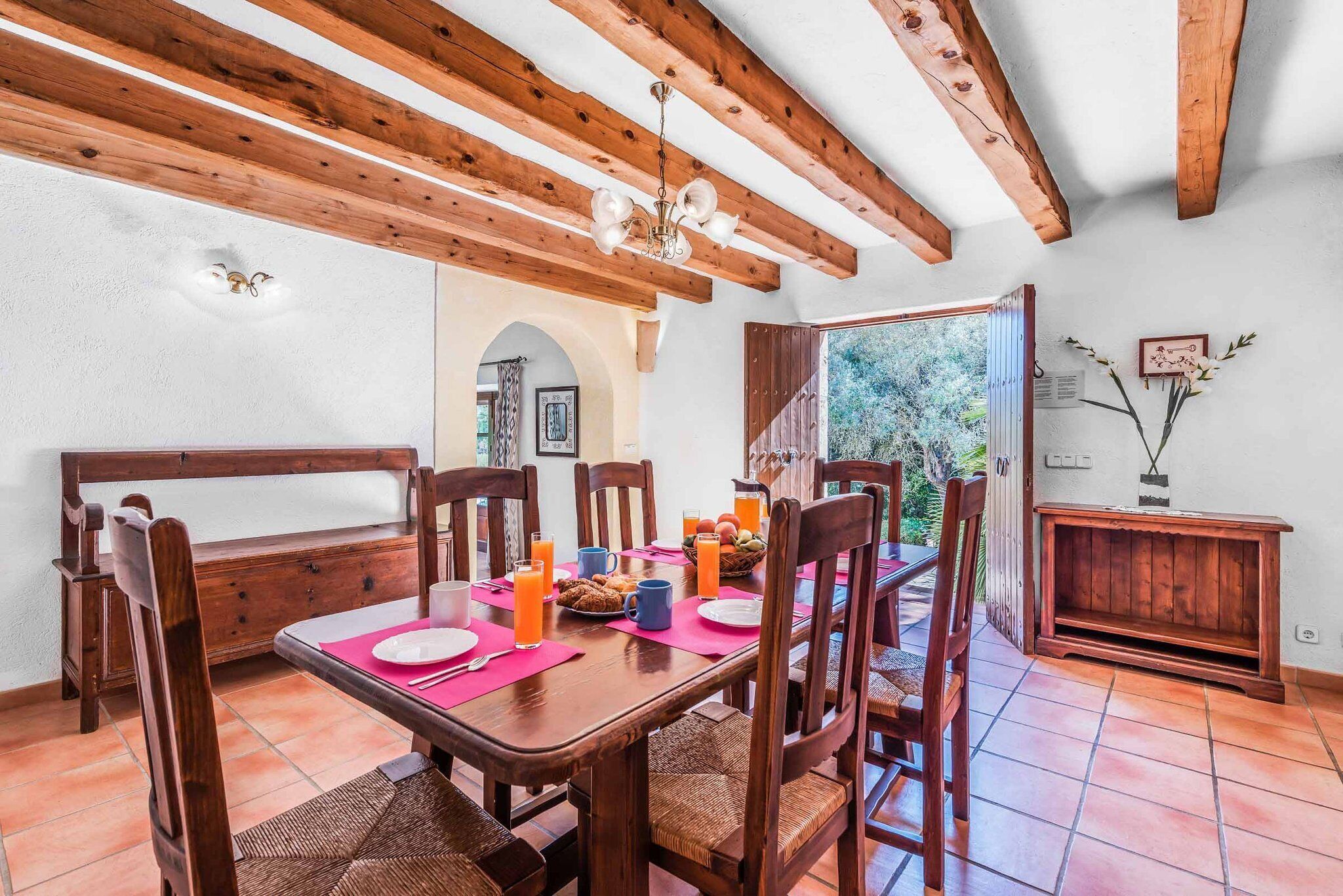 Un comedor con muebles de madera, una mesa dispuesta con platos, tazas y una cesta de fruta.