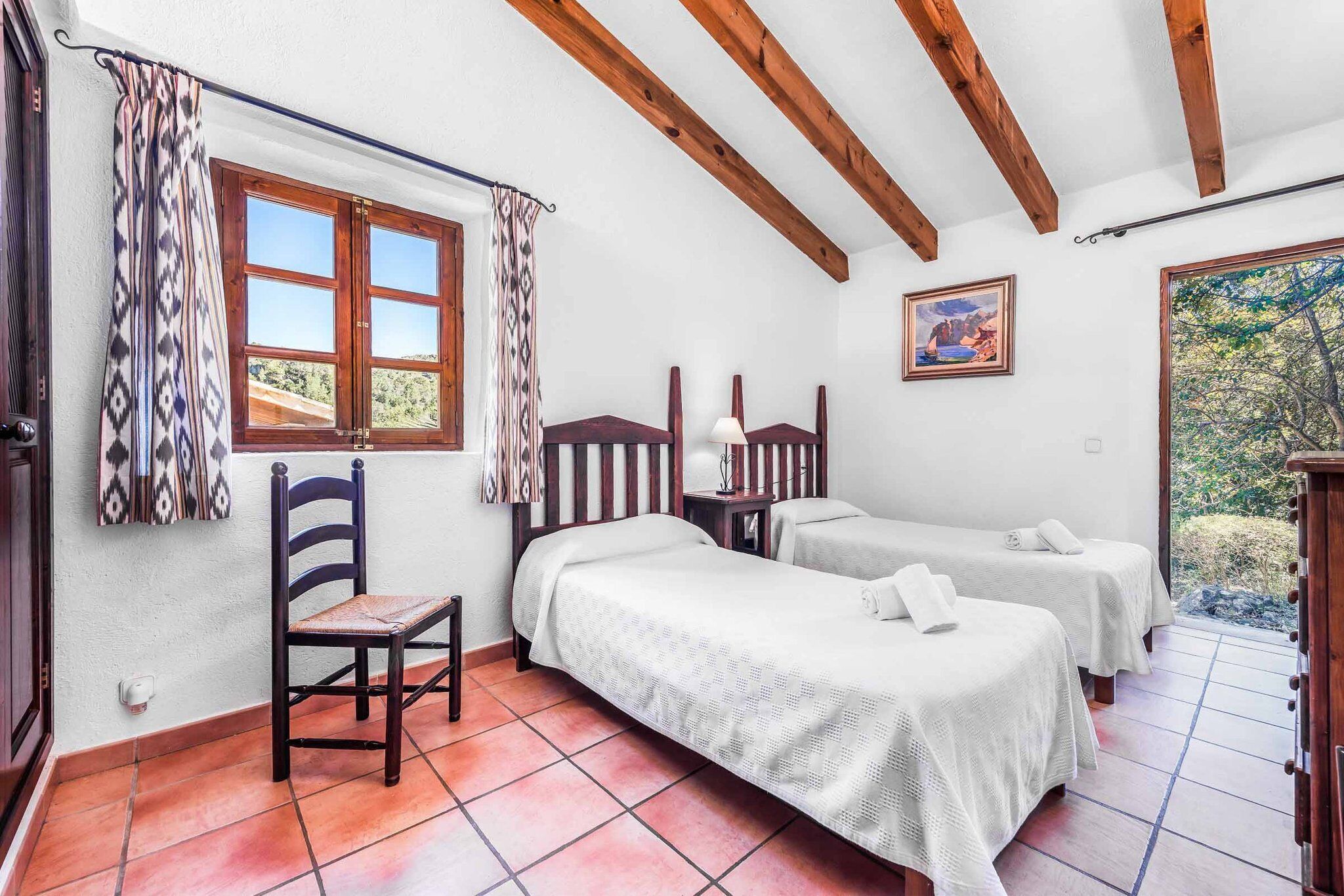 Quarto com duas camas, uma cadeira, janela com cortinas, um quadro e vista para vegetação.
