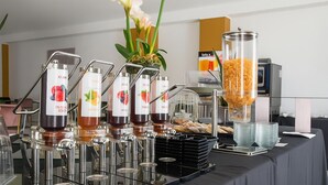 Disposición de un bufé con varios dispensadores de bebidas etiquetados con distintos sabores, como 'FRUIT CREAM' y 'NARANJA'. También hay recipientes de vidrio transparentes que parecen contener cereales o aperitivos.