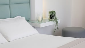 Una cama bien hecha con sábanas blancas, un cabecero tapizado en color aguamarina, un candelabro de cristal transparente y una pequeña planta en maceta sobre una mesita de noche blanca.