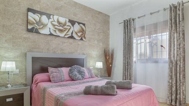 Un dormitorio con cama, almohadas, una mesita de noche, lámpara, ventana con cortinas y una obra de arte floral en la pared.
