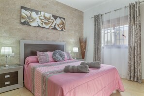 A bedroom with a bed, pillows, a nightstand, a lamp, a window with curtains, and a floral artwork on the wall. - Holiday Home 'La Casita De Leonardo' with Private Pool, Wi-Fi & Air Conditioning (Conil de la Frontera)