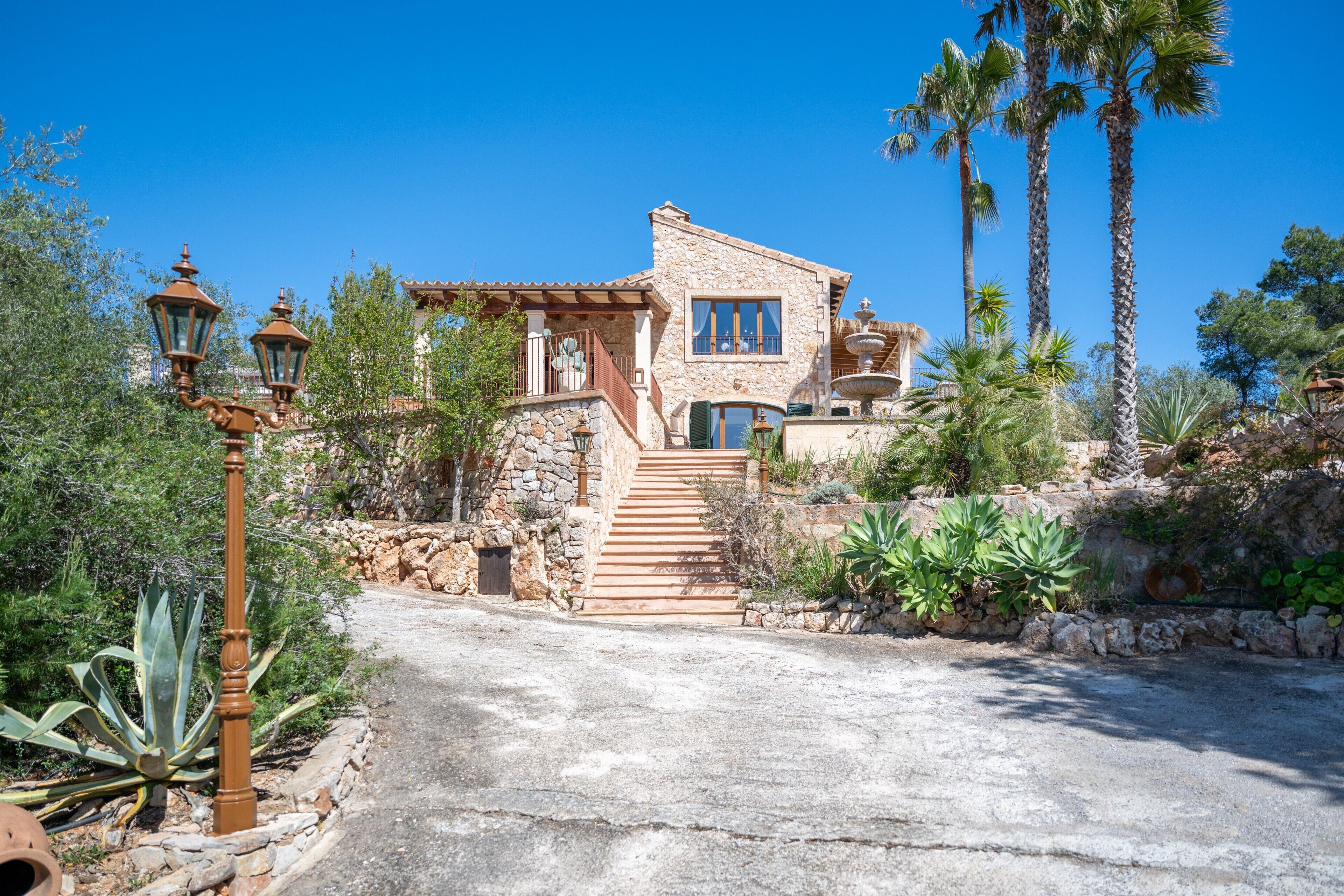 Une maison en pierre avec un escalier en pierre menant à un balcon, le tout entouré de palmiers et de cactus.