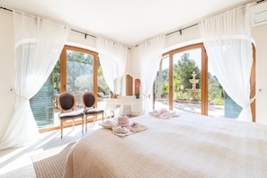 A bedroom with a large bed, a vanity, two chairs, and a view of a garden through a glass door. - Casa rural 'Vall d'Or' con vistas al mar, piscina privada y Wi-Fi (Illes Balears)
