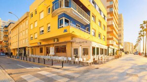 Un edificio amarillo con una cafetería llamada 'Charlotte' en la planta baja, mesas al aire libre y una calle peatonal con palmeras.