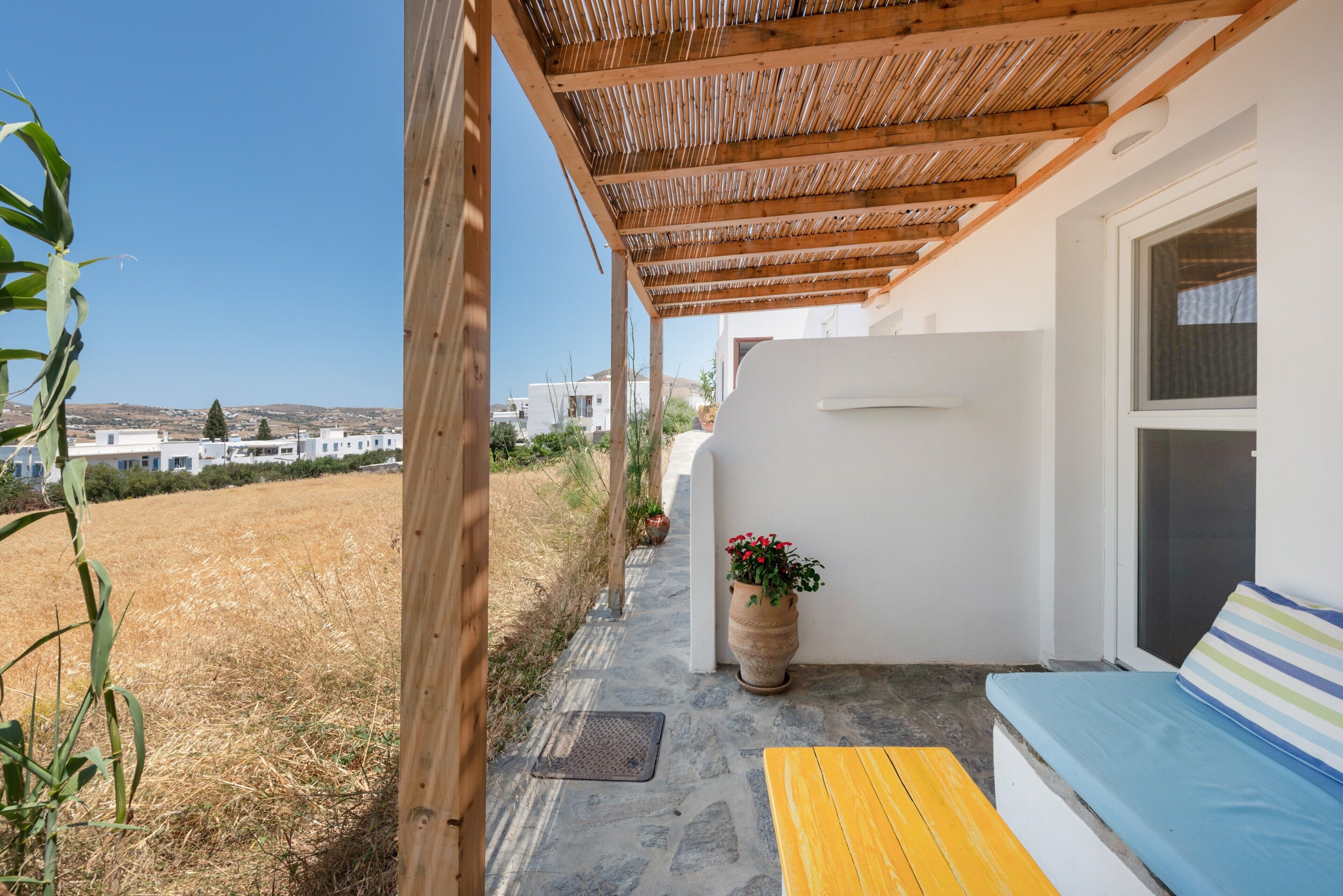A covered outdoor seating area with a stone floor, a wooden bench, a sofa with a striped cushion, and a potted plant.