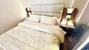 Een bed met een gestoffeerd hoofdbord, witte beddengoed en nachtkastjes met lampen en planten.
