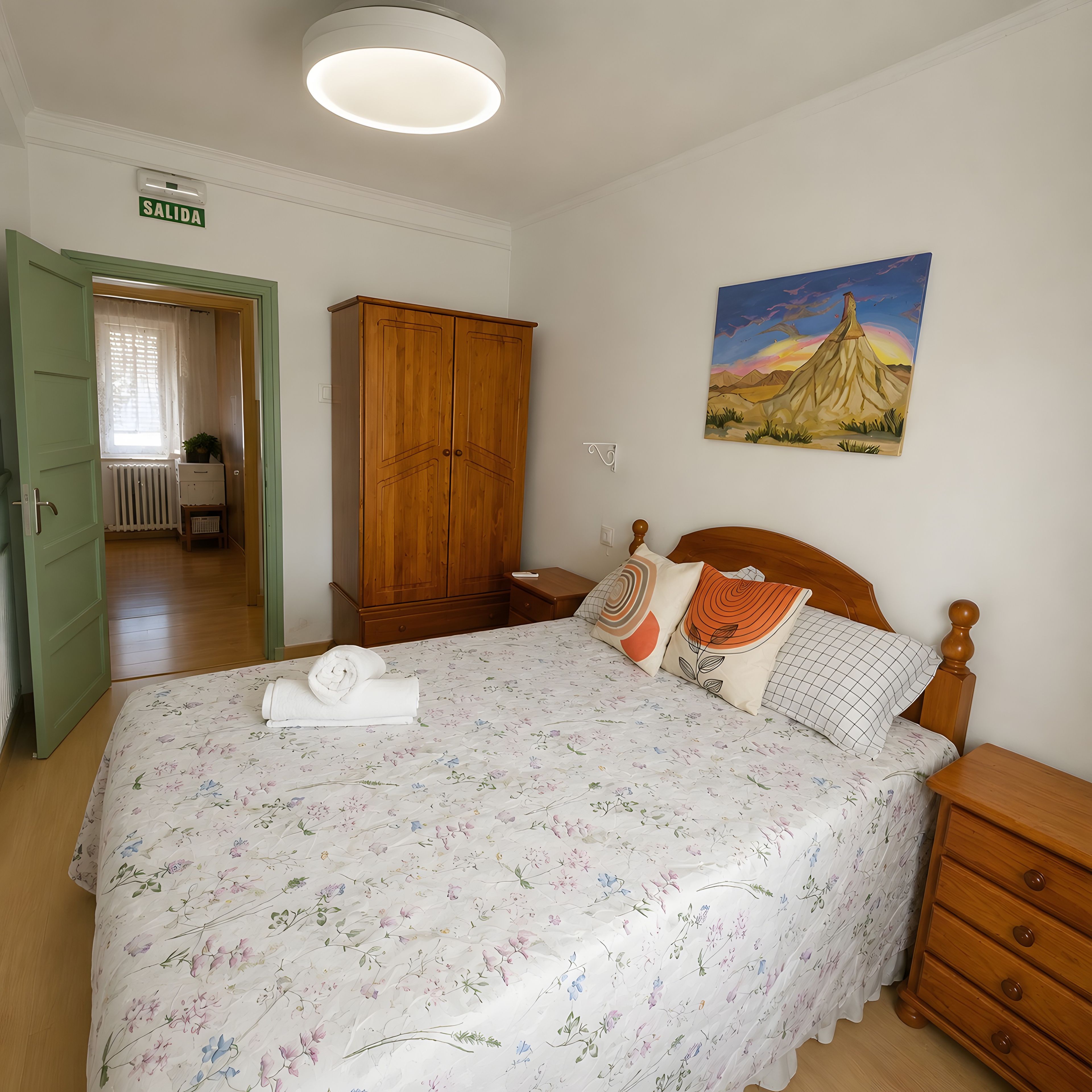 Un dormitorio con una cama, un armario de madera, un cuadro y una puerta con la etiqueta 'SALIDA'.