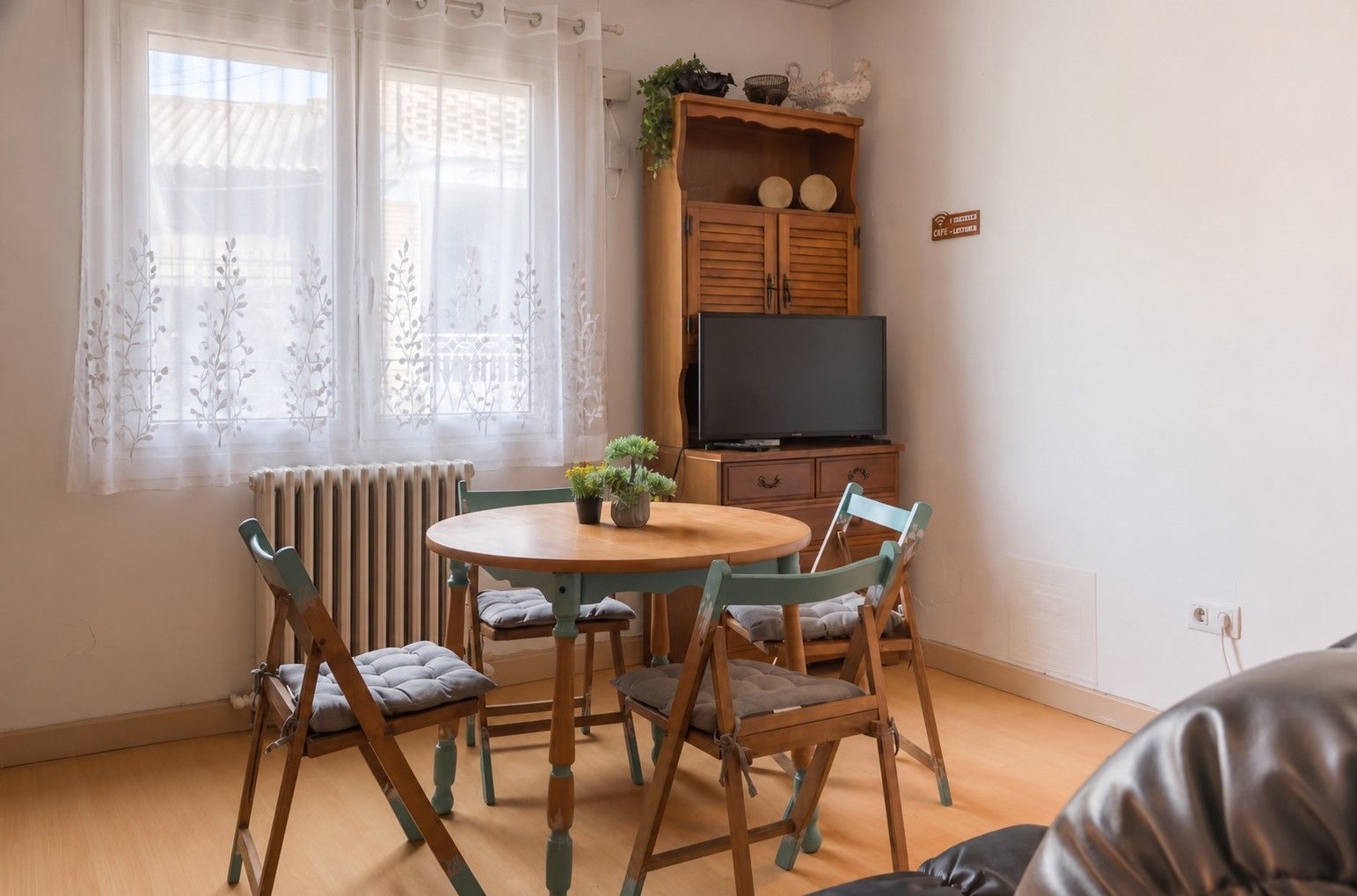 Una habitación con una mesa redonda de madera, cuatro sillas, un radiador, un televisor sobre un mueble de madera y una ventana con cortinas de gasa.