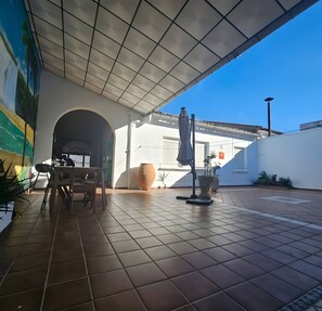 Área de varanda coberta com piso de azulejos, mesa, cadeiras e plantas em vasos.