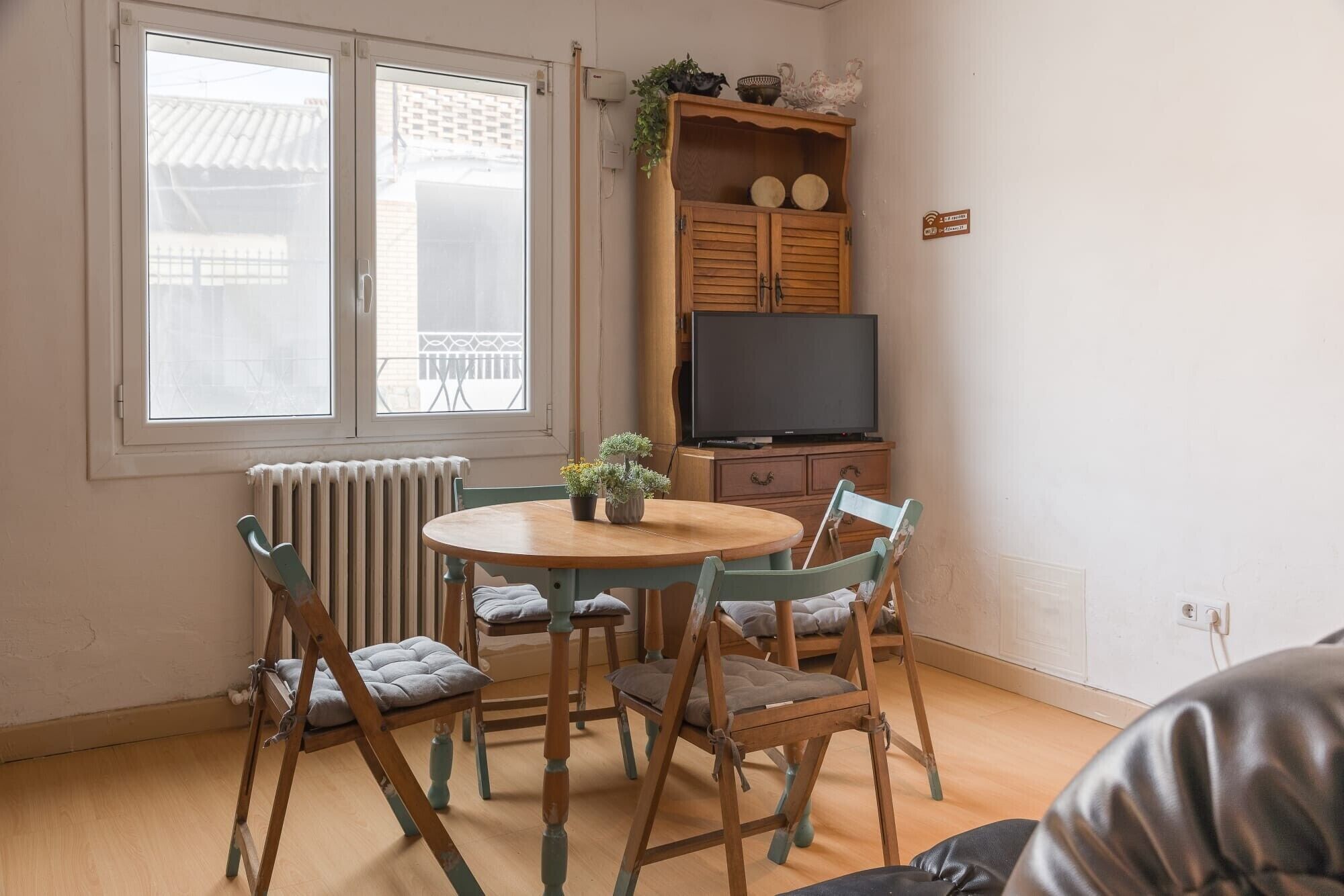 A room with a round wooden table, four chairs, a television on a wooden cabinet, and a window with a view of buildings.