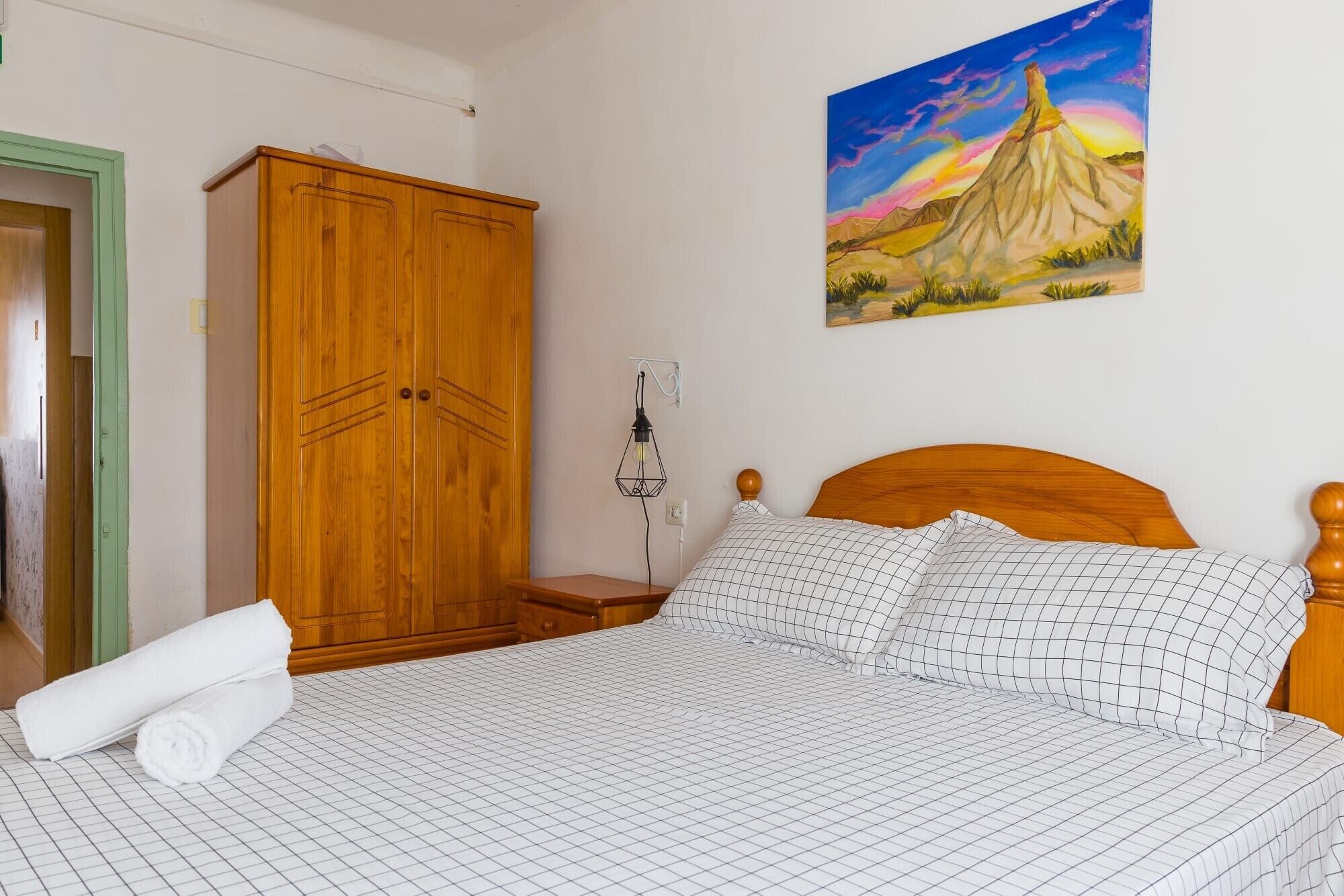 A bedroom with a wooden wardrobe, a bed with a checkered bedspread, a painting of a mountain landscape, and a wall-mounted lamp.