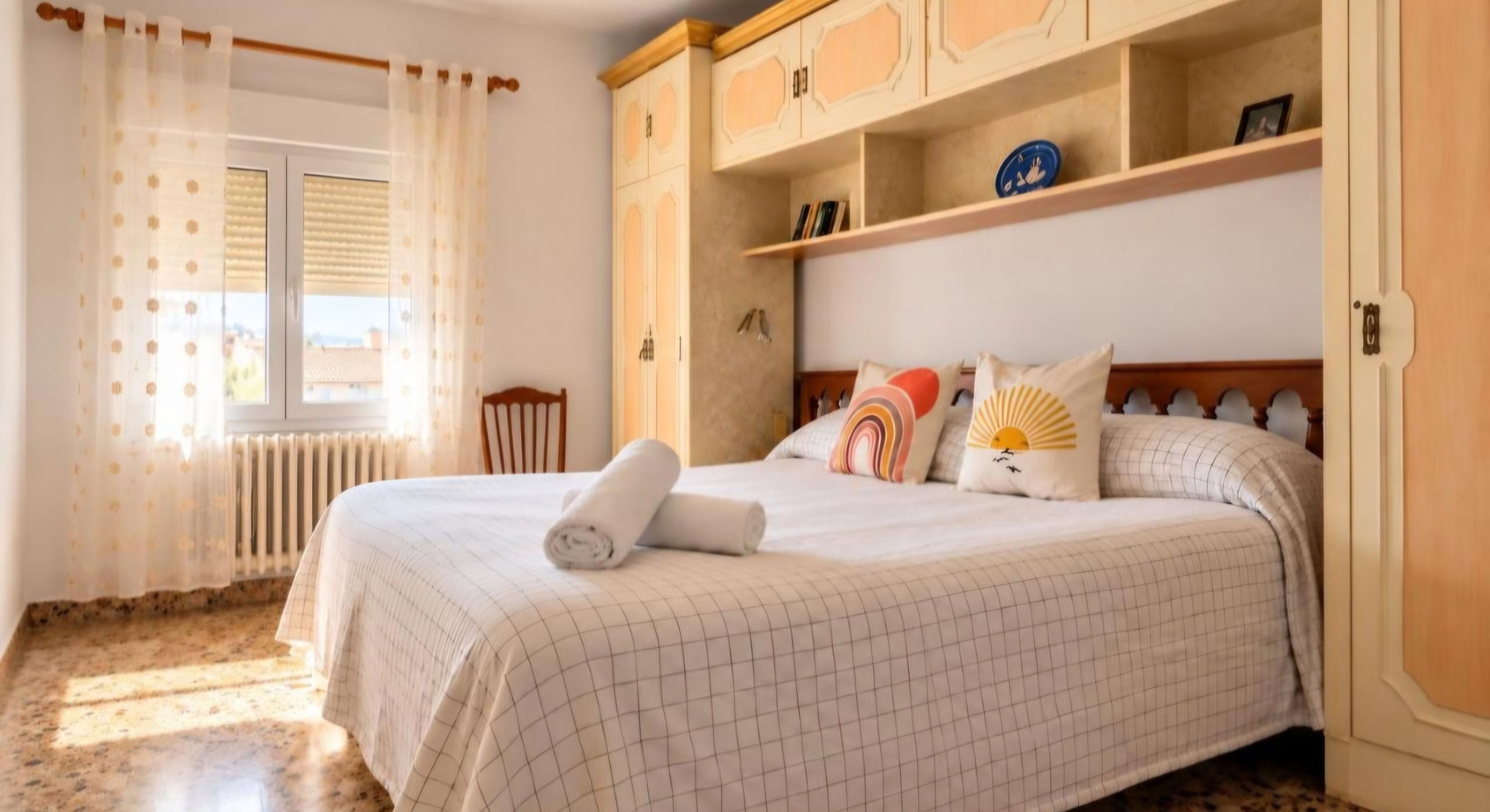 Una cama bien hecha con una toalla enrollada, cabecera de madera y una ventana con cortinas.