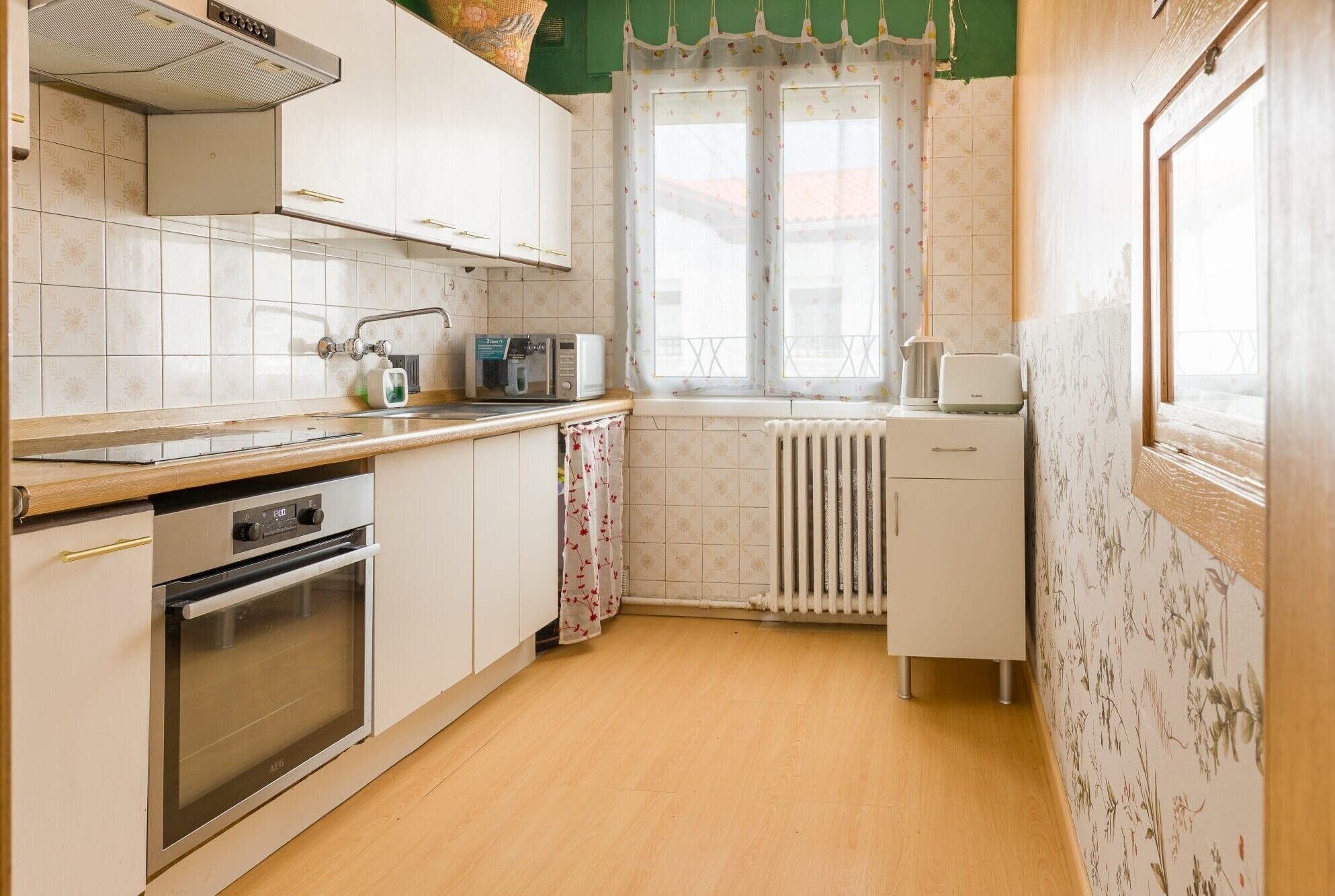 Cocina compacta con armarios blancos, horno empotrado, microondas y ventana con cortinas florales.
