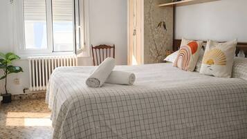 Un dormitorio con cama, radiador, ventana con persianas, una silla de madera y un armario con objetos decorativos.