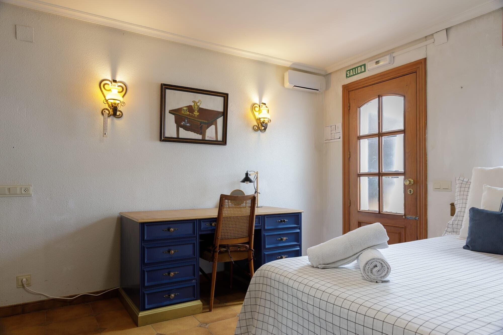 Habitació amb llit, escriptori amb calaixos, cadira, llums penjades a la paret, aparell d'aire condicionat i porta amb finestra.