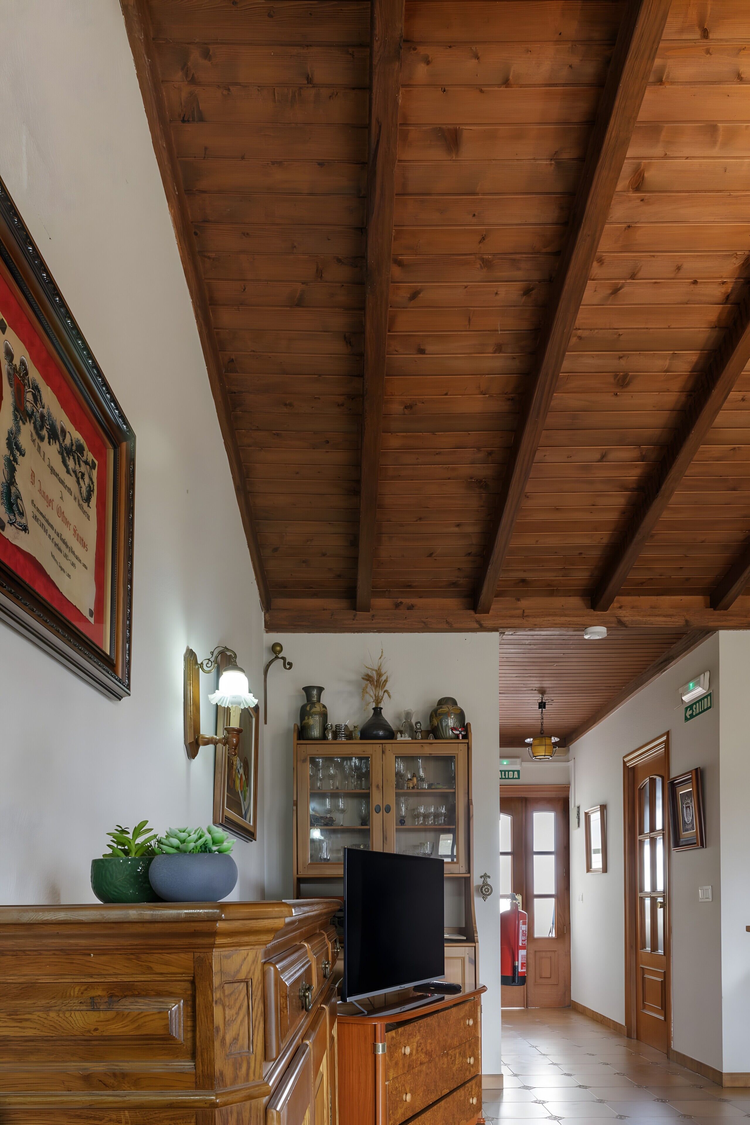 Techo de madera, un televisor sobre un mueble de madera, un certificado enmarcado en la pared y un pasillo que conduce a una puerta.
