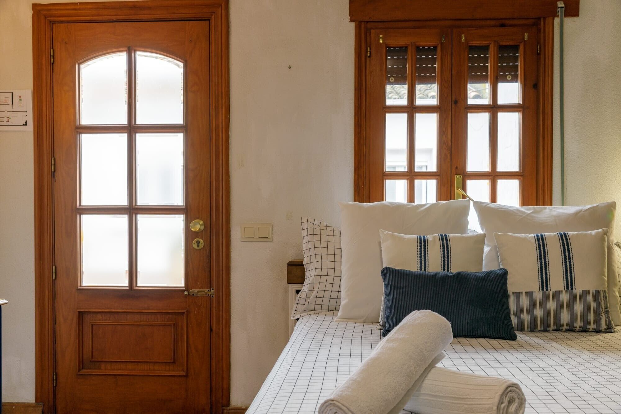 Una habitación con puerta de madera, una cama con muchos almohadones y una ventana con persianas.