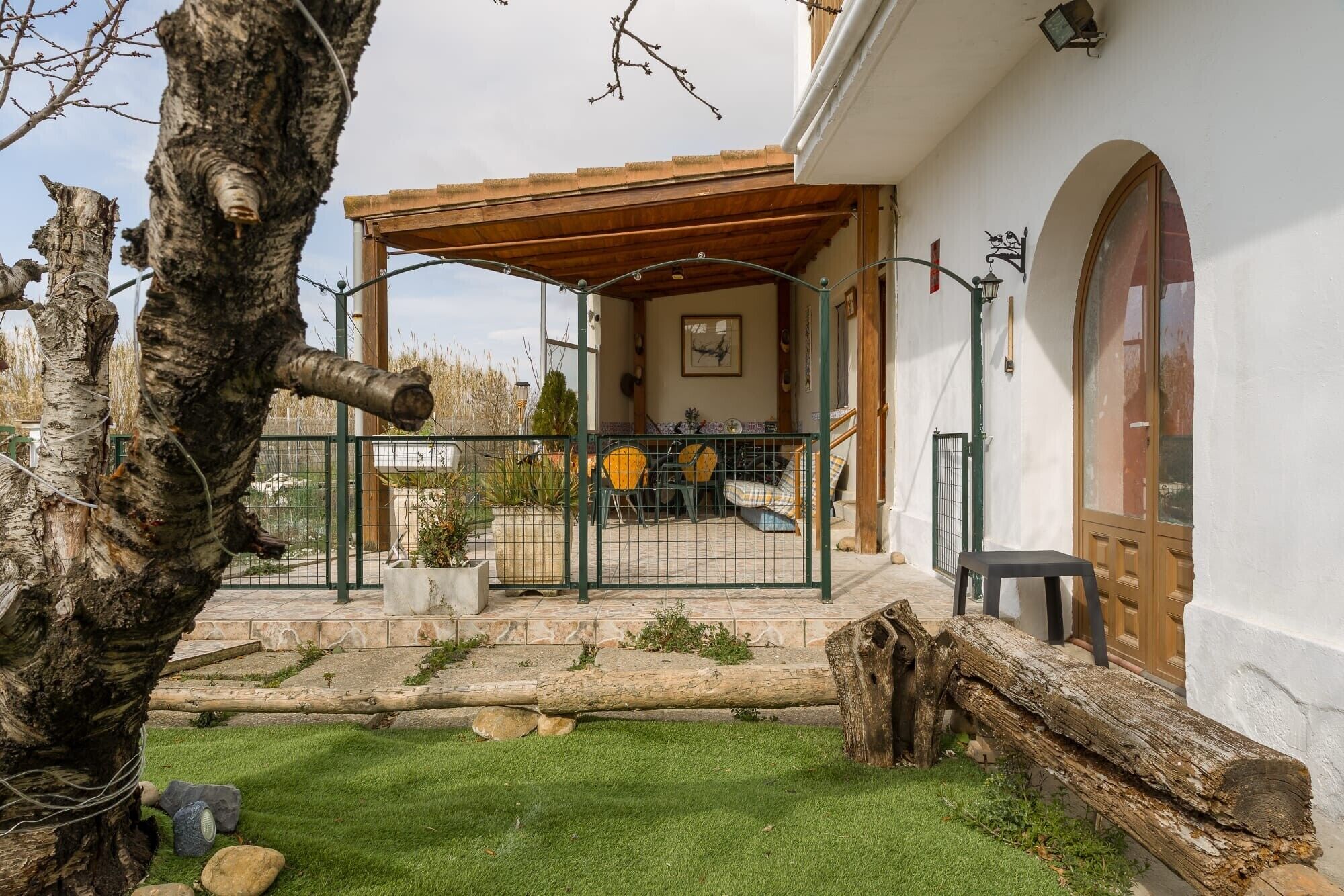 Zona d'estar a l'aire lliure coberta, amb taula i cadires, una pérgola de fusta i un edifici blanc amb una finestra d'arc.