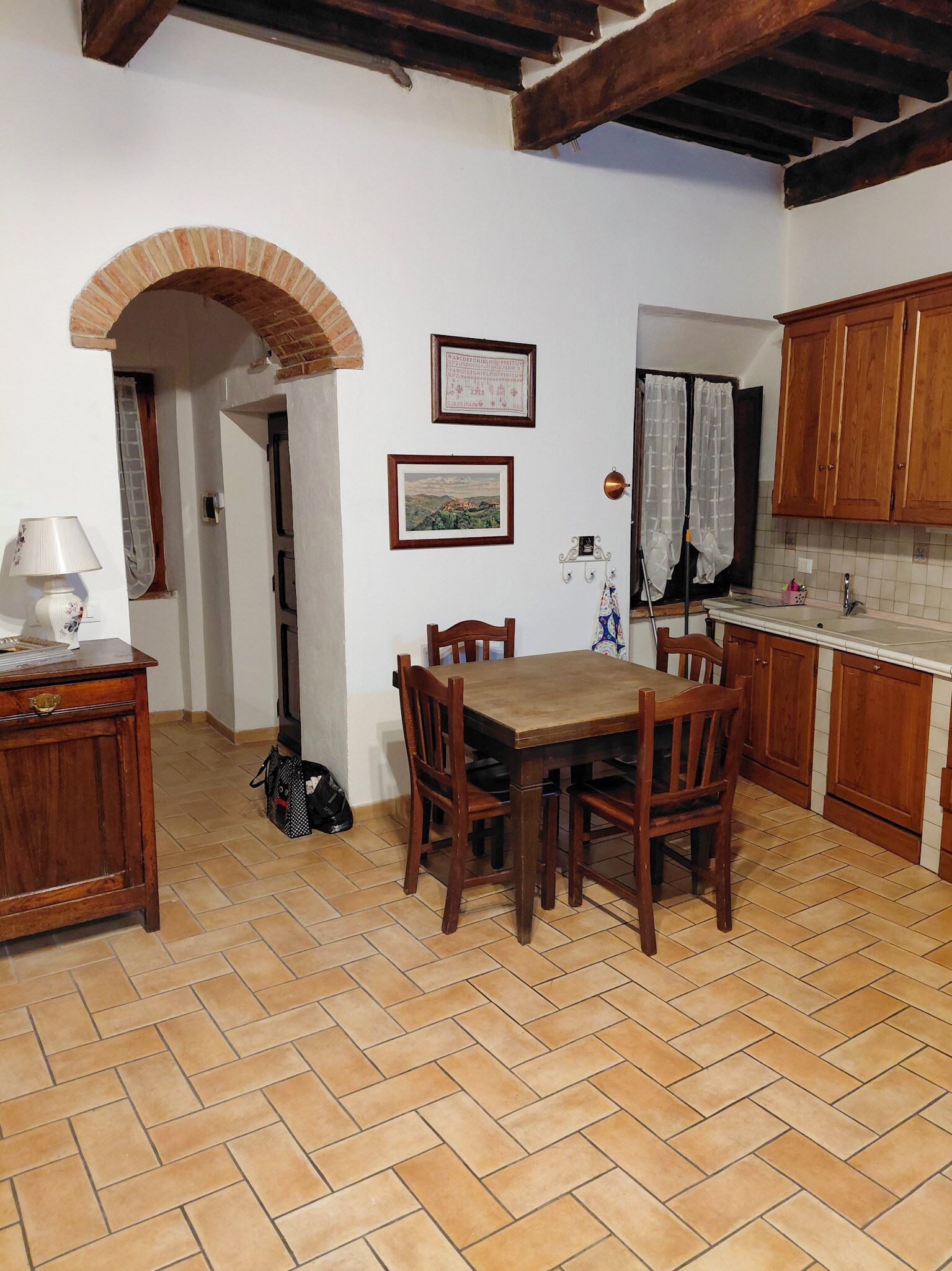 A kitchen with wooden cabinets, a tiled floor, a dining table with chairs, and a small archway leading to another room.