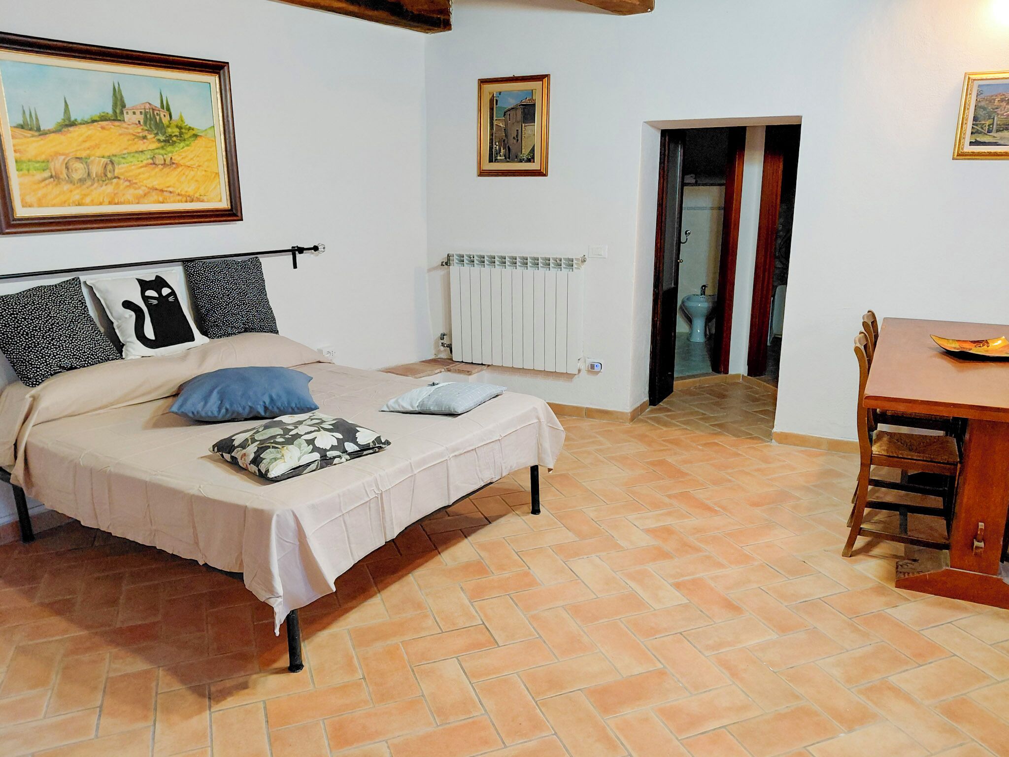 Una camera da letto con un letto, un tavolo e delle sedie. Alle pareti ci sono dei quadri e un termosifone.