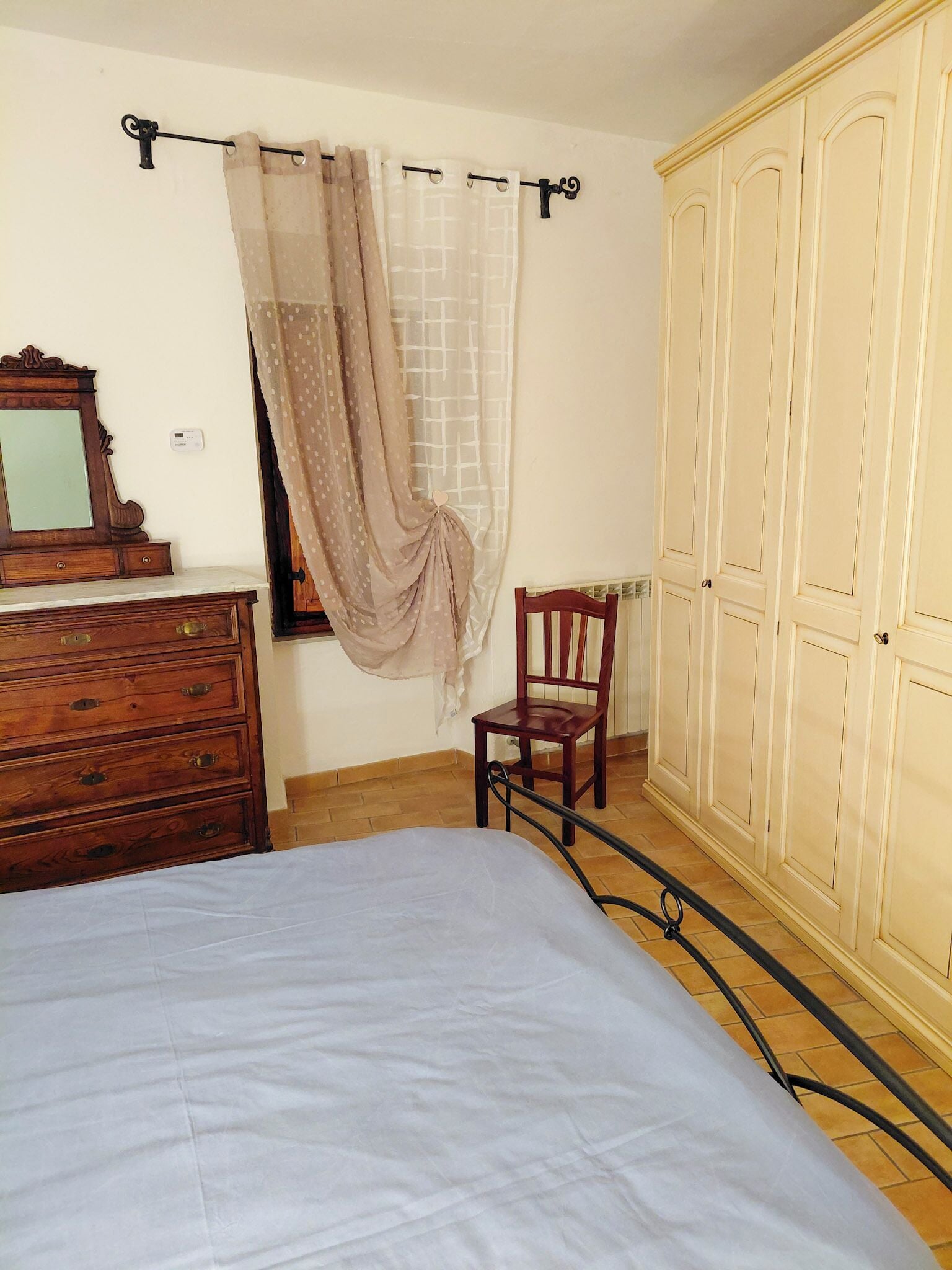 Ein Schlafzimmer mit Bett, Holzkommode, Stuhl, Kleiderschrank und einem Fenster mit Vorhängen.