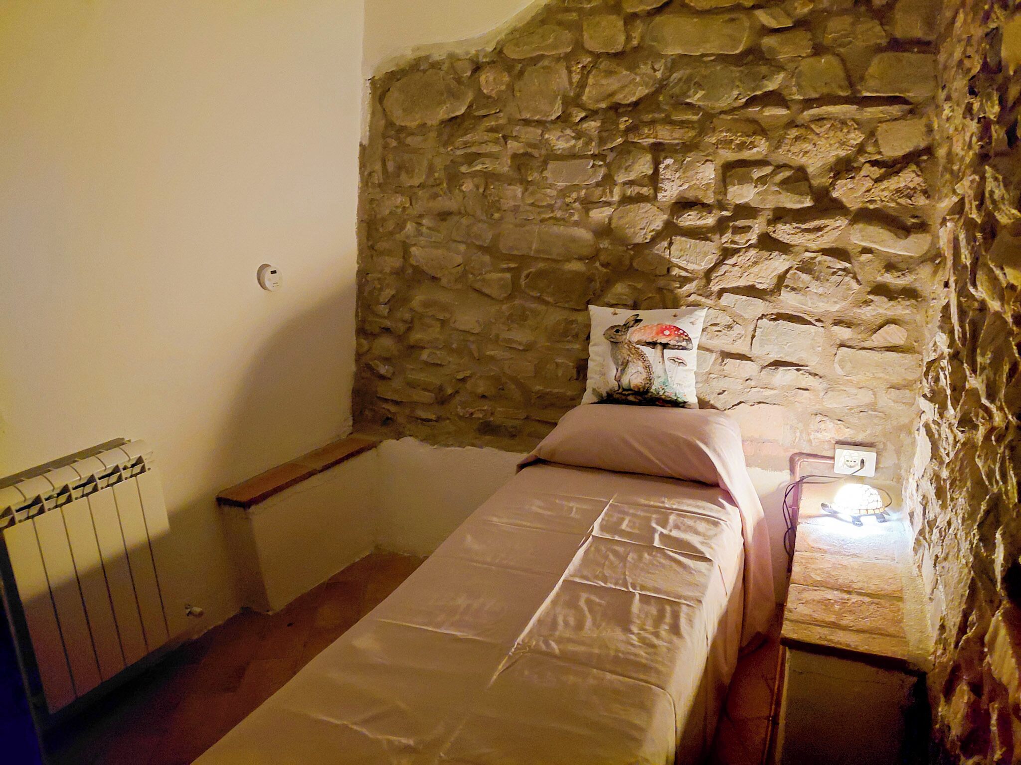 A small, stone-walled room with a single bed, a nightstand, and a radiator.