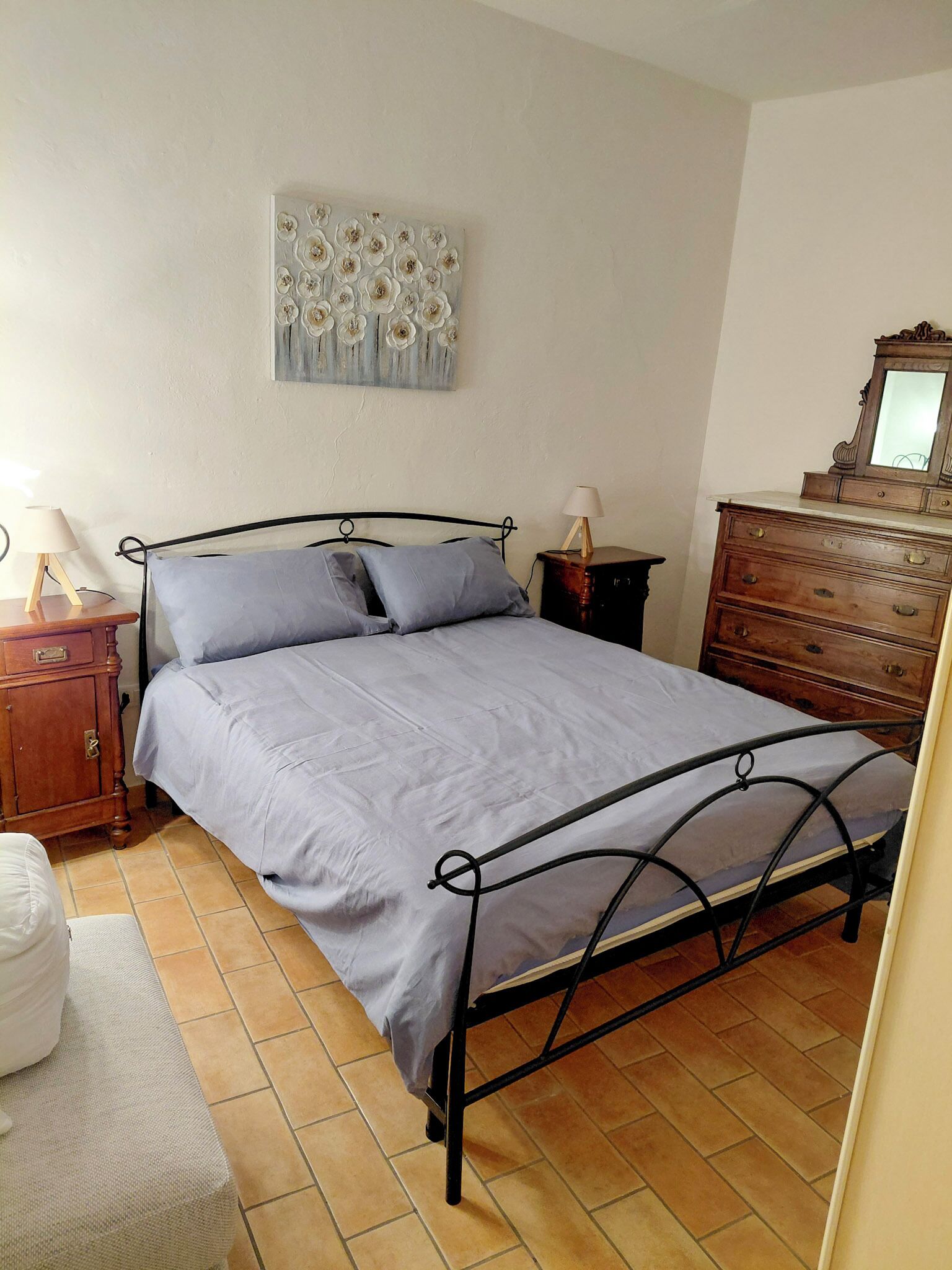 Een slaapkamer met een metalen bed, een houten dressoir en een spiegel aan de muur.