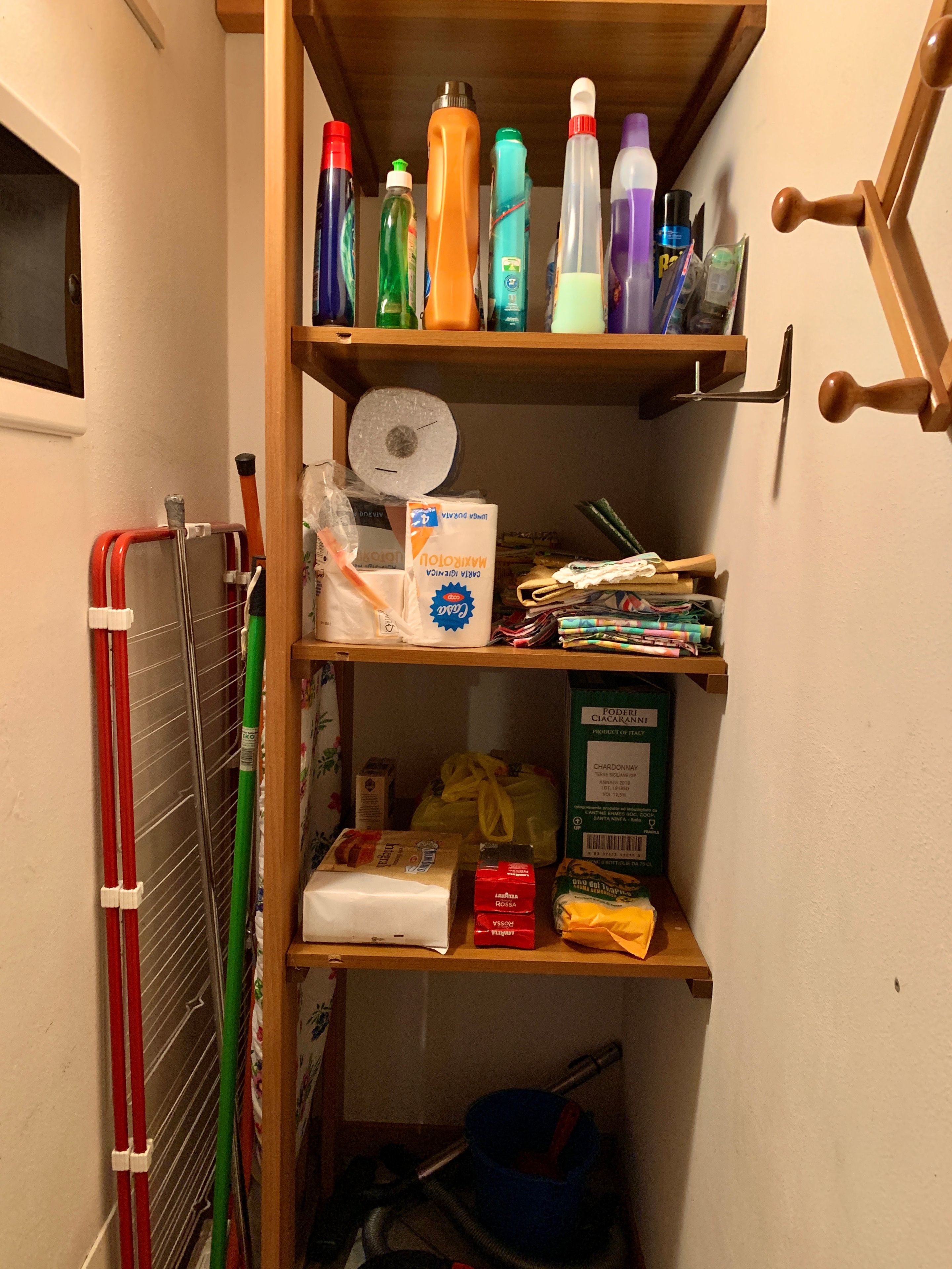 A narrow storage space with shelves holding various items including cleaning supplies, a vacuum cleaner, and a small box.