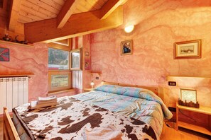 A bedroom with a bed, a nightstand, a lamp, a window, and a wall with framed pictures. - Casa Aliza – Alpine Duplex in the Heart of Valle del Chiese (Creto)