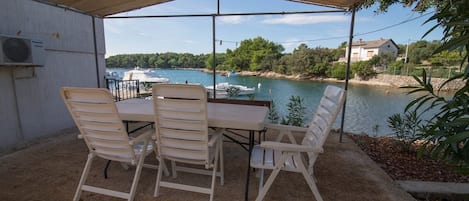 Een overdekt terras met een tafel en stoelen, met uitzicht op een waterpartij met boten en huizen op de achtergrond.