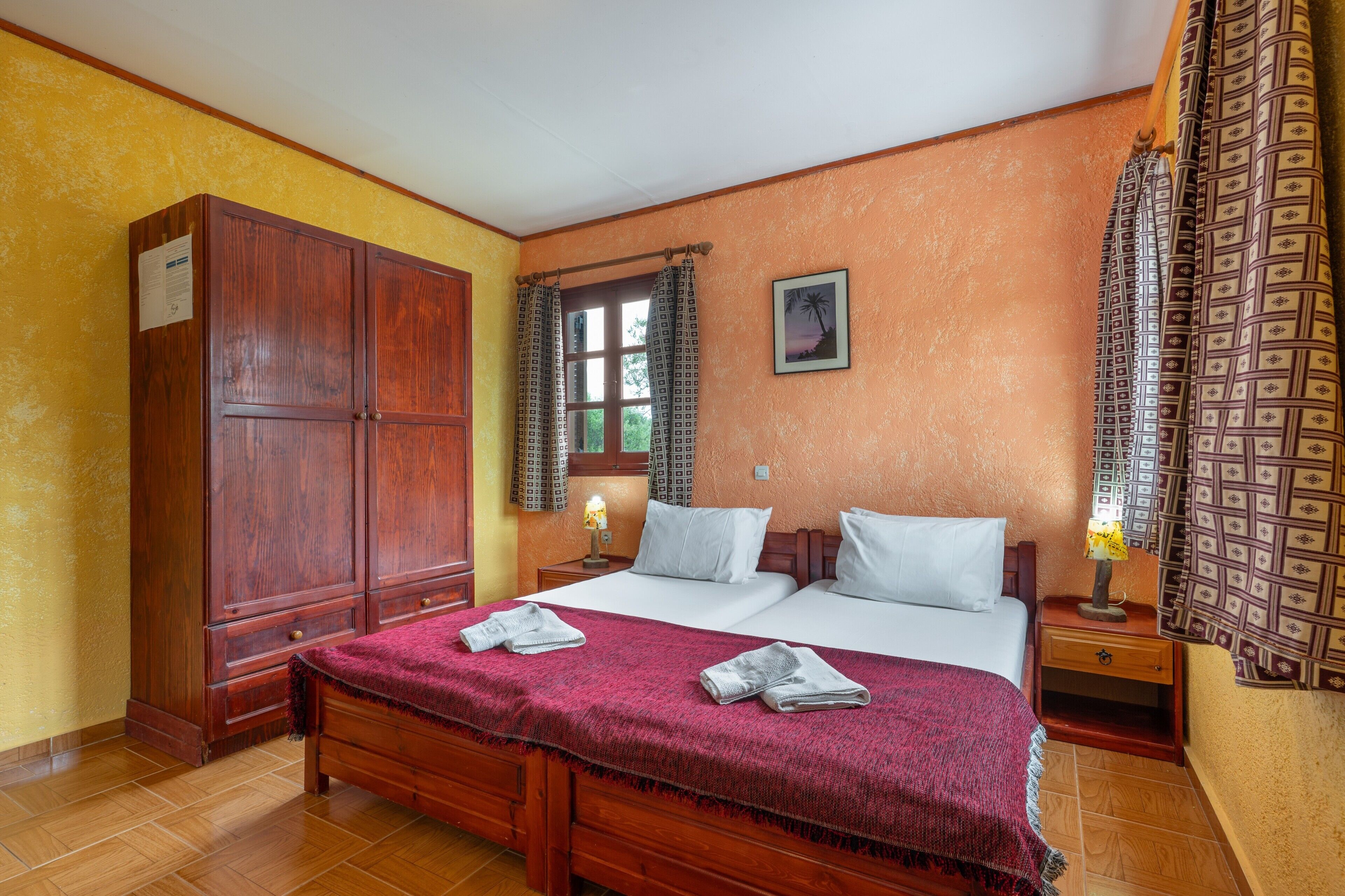 A bedroom with a wooden wardrobe, a bed with white linens, a nightstand, and a window with patterned curtains.