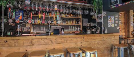 A bar with a wooden counter, metal stools, and a variety of bottles and glasses.