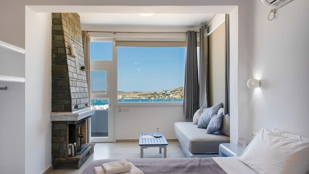 A bedroom with a stone fireplace, a bed, a sofa, a small table, and a balcony with a view of the sea. - Holiday Home 'Saphhire' with Sea View, Shared Terrace and Wi-Fi (Paros)