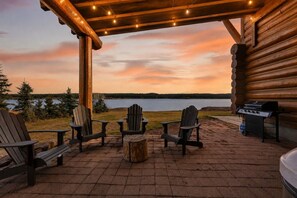 Una cabaña de troncos con un patio, mobiliario exterior, una parrilla y vistas a un lago al atardecer.