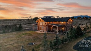 A log cabin with a covered porch and a hot tub, surrounded by a natural landscape and mountains in the background. - Stone Skipper | Waterfront! Private Dock! Boat Ramp! Hot Tub! (Island Park)