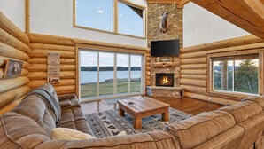 A log cabin interior with a stone fireplace, a large window overlooking a body of water, and a cozy seating area. - Stone Skipper | Waterfront! Private Dock! Boat Ramp! Hot Tub! (Island Park)