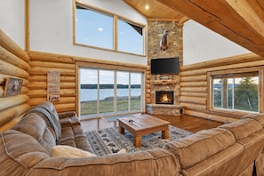 L’intérieur d’une cabane en rondins, avec une cheminée en pierre, une grande fenêtre donnant sur un plan d’eau et un coin salon confortable.