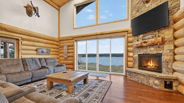 A log cabin interior with a fireplace, a sofa, a coffee table, and a large window overlooking a lake. - Stone Skipper | Waterfront! Private Dock! Boat Ramp! Hot Tub! (Island Park)