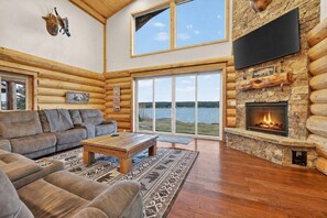 A log cabin interior with a fireplace, a sofa, a coffee table, and a large window overlooking a lake. - Stone Skipper | Waterfront! Private Dock! Boat Ramp! Hot Tub! (Island Park)