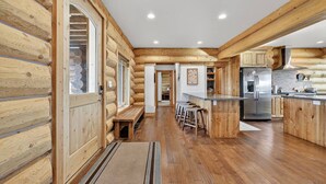 A log cabin interior with a kitchen area featuring wooden cabinetry, a stainless steel refrigerator, and a bar area with stools. - Stone Skipper | Waterfront! Private Dock! Boat Ramp! Hot Tub! (Island Park)