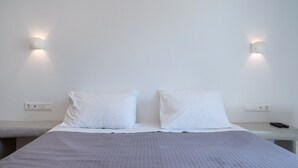 Un lit recouvert d’une courtepointe matelassée, deux oreillers blancs, une table de chevet avec une télécommande et des lampes fixées au mur.