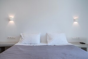 Un lit recouvert d’une courtepointe matelassée, deux oreillers blancs, une table de chevet avec une télécommande et des lampes fixées au mur.