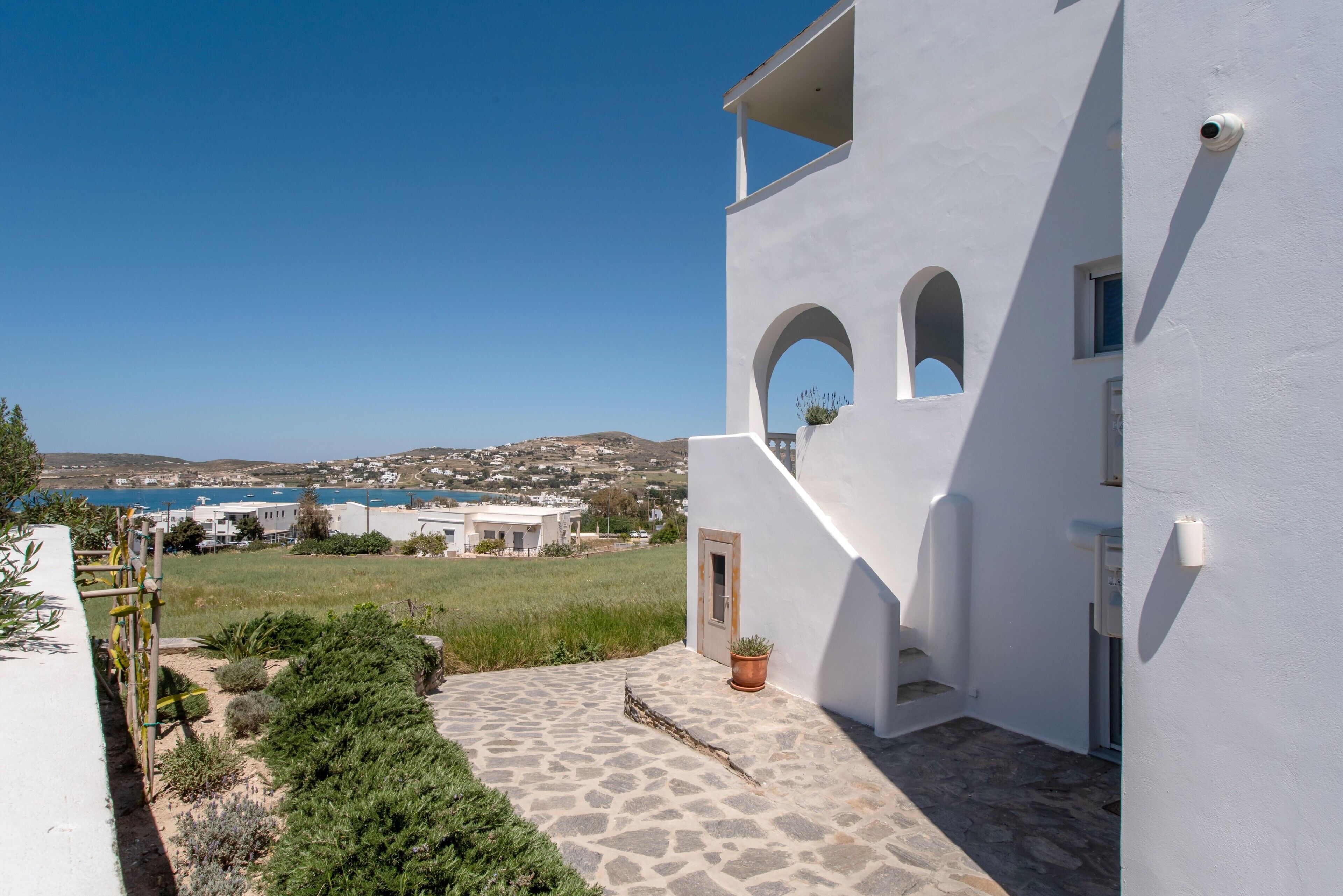 A white building with a stone pathway, a potted plant, and a view of the sea and hills.
